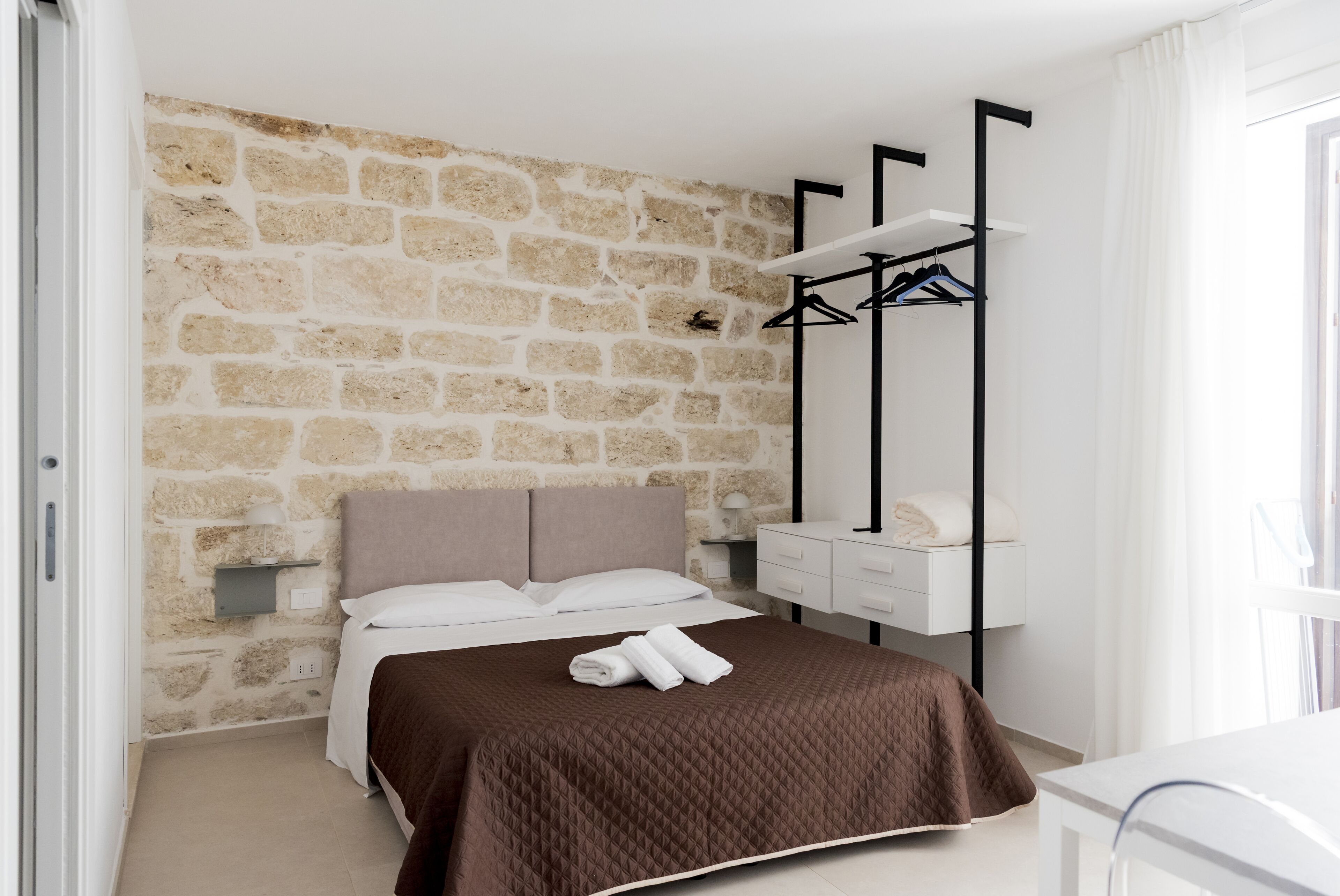 A bedroom with a stone wall, a bed with a brown quilt, a bedside table with a lamp, and a clothing rack.