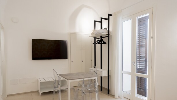 A room with a bed, a TV mounted on the wall, a table with chairs, a coat rack, and a door with blinds. - Sky D'Alcamo Ruby (Alcamo)