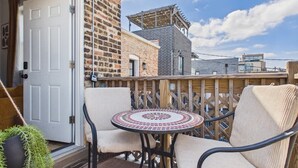 A balcony with wicker chairs and a round table, a wooden fence, and a brick building in the background.