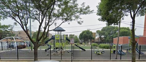 Un parque infantil con una estructura para escalar, un área cercada, un coche estacionado y árboles.