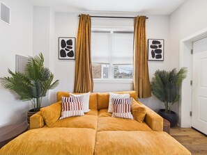 Una habitación con un sofá grande, anaranjado y con capitonado, dos plantas en macetas y una ventana con cortinas.