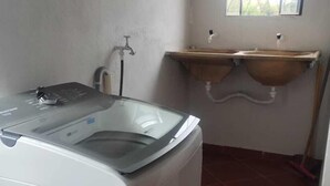 A compact laundry room with a washing machine, a window with star-shaped bars, and a small sink with a wooden countertop. - Confort Suite 01 (Contagem)