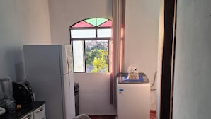 A compact kitchen with a refrigerator, a coffee maker, a microwave, and a small sink. There is a window with a view of buildings and trees outside. - Confort Suite 01 (Contagem)