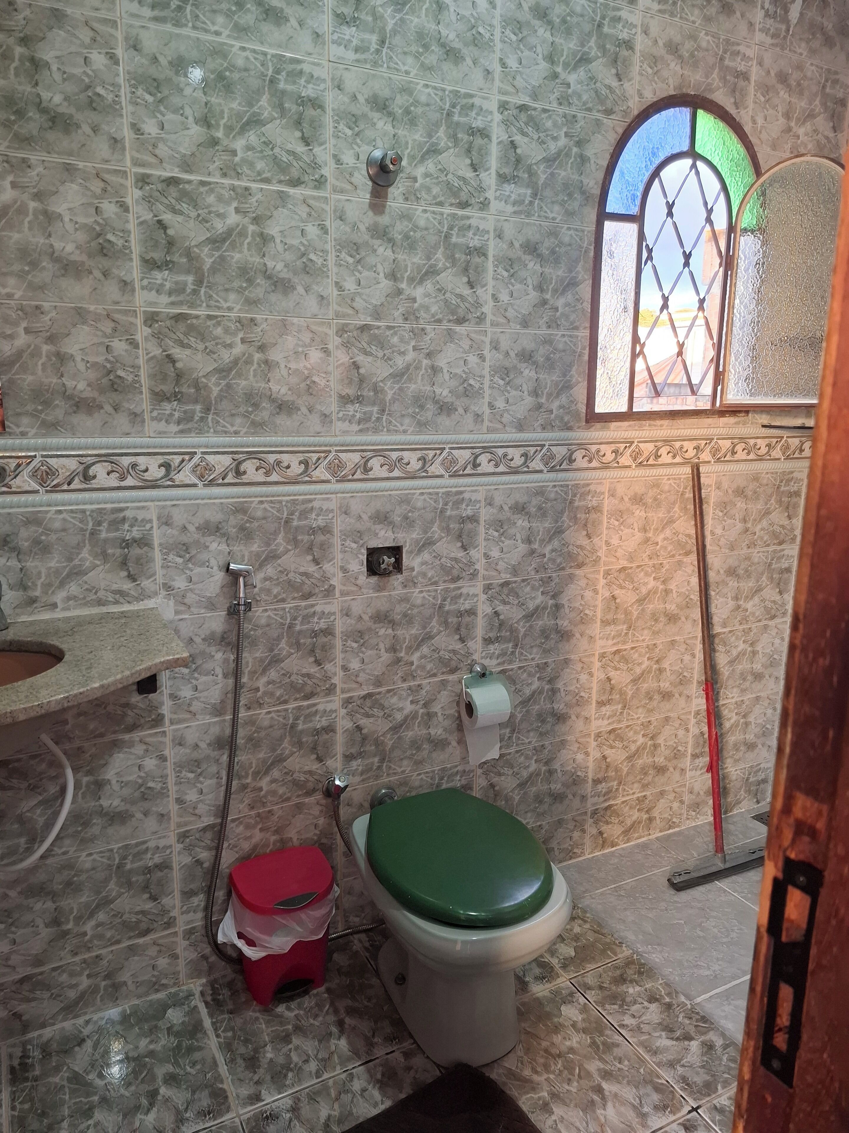 Un baño con baldosas de mármol, un inodoro con asiento verde, un cubo de basura rojo y una ventana con vistas al exterior.