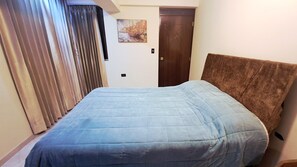 A hotel room with a large bed, a window with curtains, a door, and a painting on the wall.