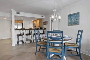 Un comedor con una mesa redonda, sillas azules y una araña. Se aprecia una cocina al fondo con armarios de madera y un microondas.