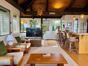 A spacious living area with a large flat-screen TV, wooden furniture, and a kitchen area with bar stools. - Hale Mahealani, Private Treetop Home, Ocean Views (Princeville)