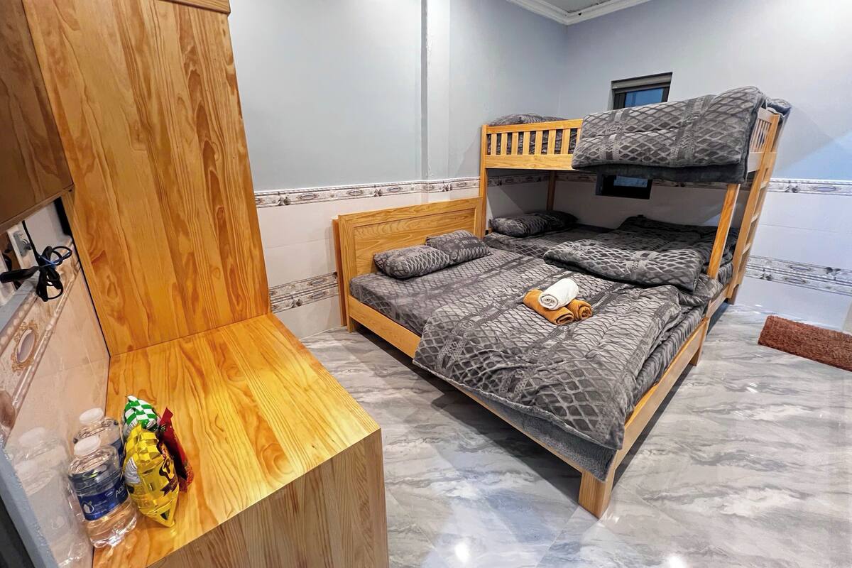 Una cama litera con cabecera de madera y una cama inferior con ropa de cama estampada. Una pequeña mesa de madera con una botella y un paquete de aperitivos está al lado de la cama.