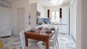 Un comedor con mesa de madera y sillas blancas, una cocina con azulejos azul y blanco, y un ventanal con vistas.