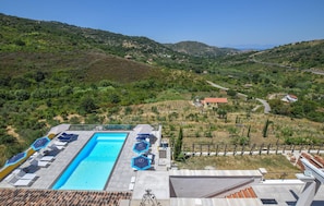 Un espace piscine doté de chaises longues et de parasols, entouré d’un paysage vallonné offrant une vue sur des maisons situées au loin.