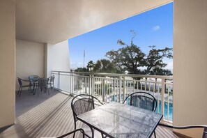 Un balcon avec une table et des chaises, une piscine en arrière-plan et une vue sur des arbres et un ciel dégagé.
