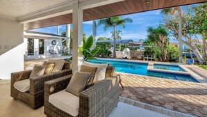 Un espace extérieur abrité, aménagé avec des meubles en osier, une piscine et offrant une vue sur une maison et des palmiers.