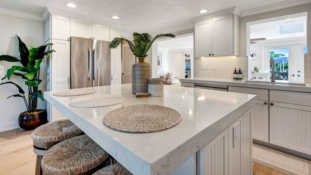 Une cuisine moderne avec des meubles blancs, un grand îlot central doté de tabourets en osier et une plante en pot.