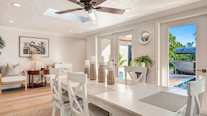 Un coin repas avec une table blanche, des chaises et un ventilateur de plafond. Une porte donne accès à un espace extérieur où se trouvent un bateau et de la verdure.