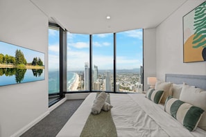 Ein modernes Hotelzimmer mit einem großen Bett, Blick auf Strand und Stadtlandschaft durch raumhohe Fenster und einem Gemälde an der Wand.