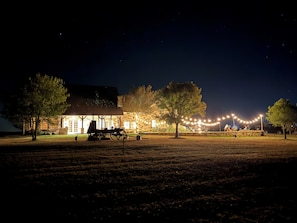 A rustic building with a stone facade, a wooden deck, and a string of lights in the foreground. - Retreat‑Ready Ranch House on 2R Ranch — Quiet, Scenic, Private (Henrietta)