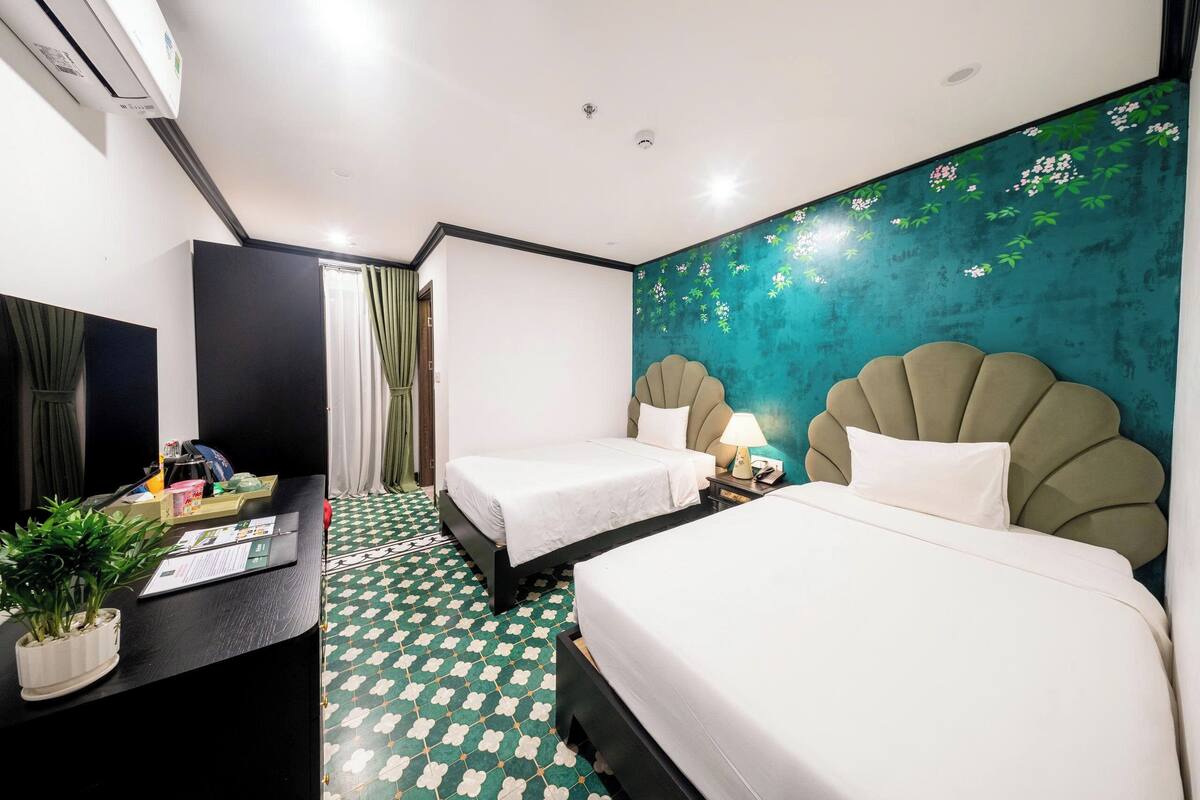 Habitación de hotel con dos camas, un escritorio con planta y pared verde azulado con estampado floral.