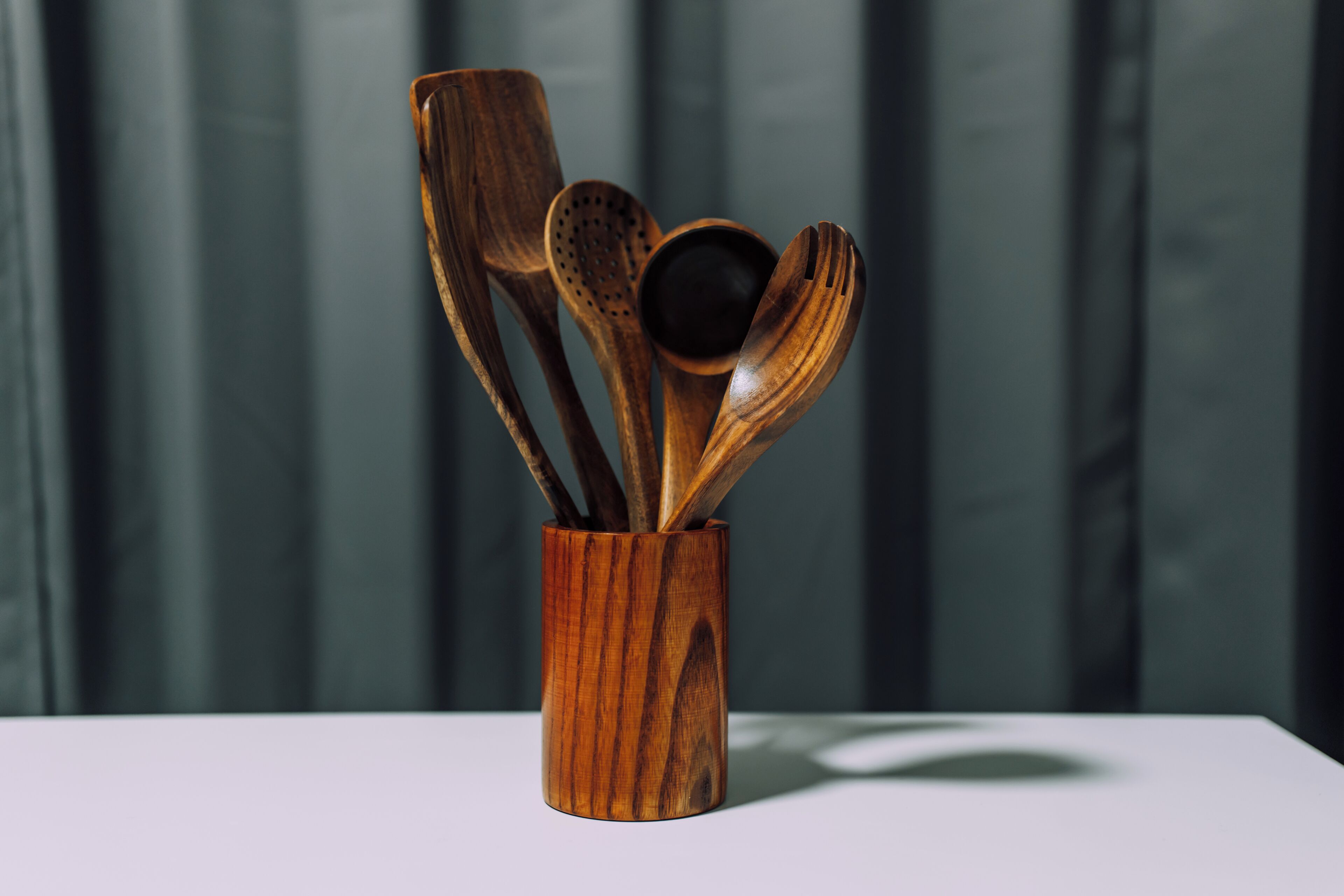 Un porte-ustensiles en bois contenant divers ustensiles en bois, posé sur une surface blanche, le tout sur un fond sombre.