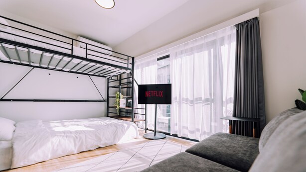 Moderni hotellihuone, jossa on kerrossänky, harmaa sohva, litteänäyttöinen televisio ja suuri ikkuna, jossa on läpikuultavat verhot.