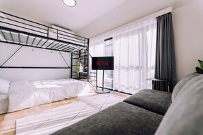 Una habitación de hotel moderna con una cama litera, un sofá gris, un televisor de pantalla plana y un ventanal con cortinas translúcidas.
