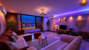 Una sala de estar moderna con un sofá grande, un televisor y vistas a un paisaje urbano nocturno.