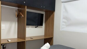 Quarto de hotel com uma cama, uma televisão presa a um móvel de madeira e uma janela com persianas.