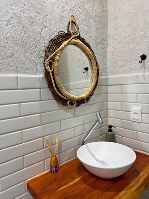 Uma casa de banho com um espelho redondo, paredes revestidas a azulejos brancos, bancada de madeira, lavatório branco, um frasco de sabonete e um difusor de vime.