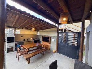 Um pátio externo com mesa e bancos de madeira, churrasqueira e televisão.