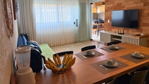 A modern living room with a dining area, a flat-screen TV mounted on a brick wall, and a large window with sheer curtains. - MHV Milagres - Mandí 03 (São Miguel dos Milagres)
