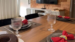 A dining area with a wooden table set for two, a television mounted on a brick wall, and a modern interior design. - MHV Milagres - Mandí 03 (São Miguel dos Milagres)