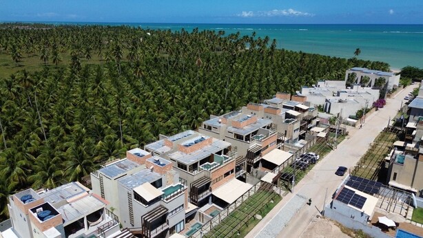 A residential area with modern buildings, a road, and a dense palm tree area in the background. - MHV Milagres - Mandí 03 (São Miguel dos Milagres)