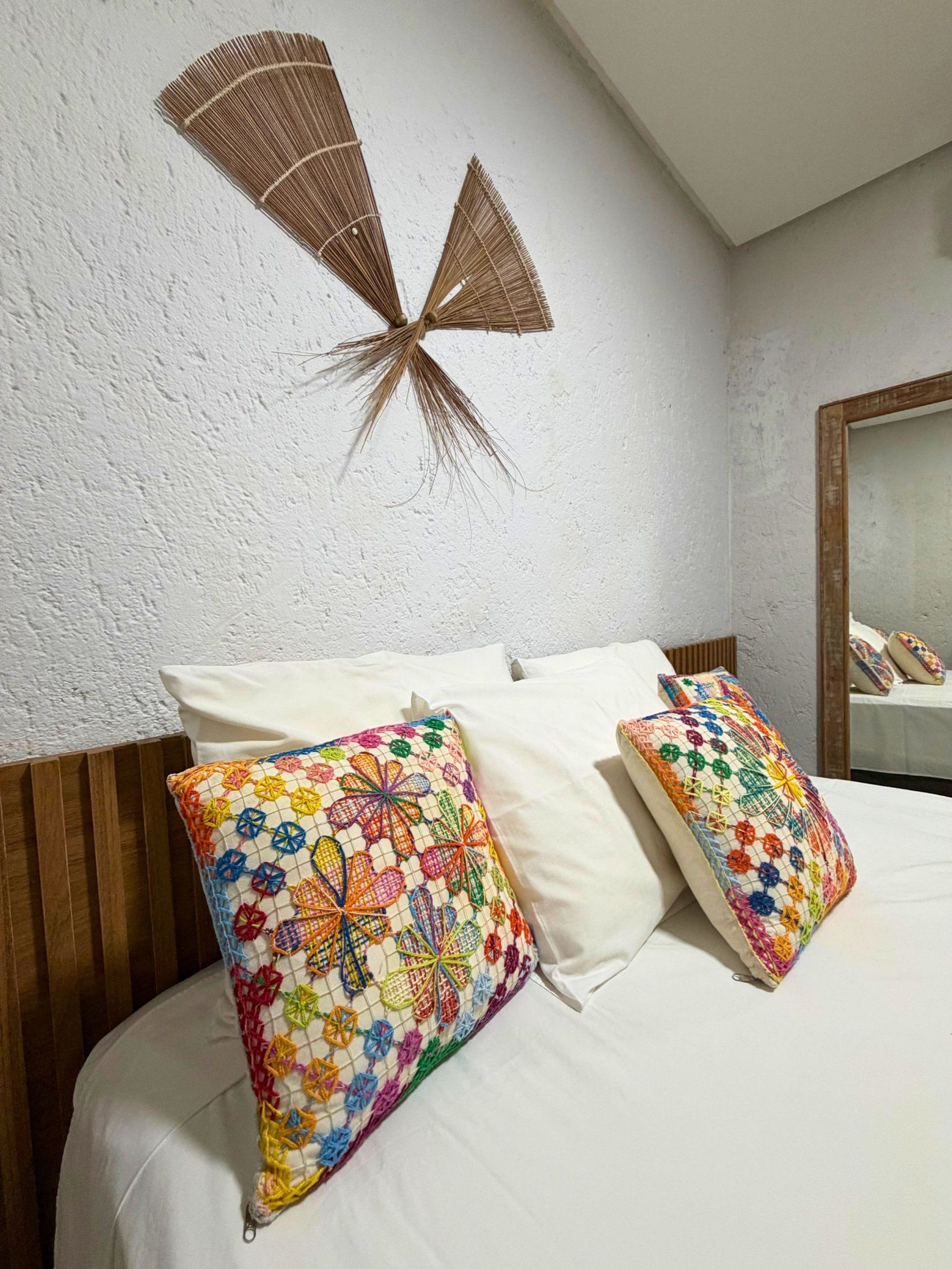 Cama com roupa de cama branca, dois travesseiros coloridos de crochê e duas ventoinhas fixadas na parede.