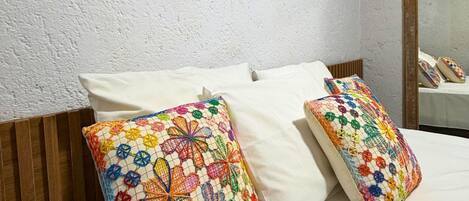 Cama com roupa de cama branca, dois travesseiros coloridos de crochê e duas ventoinhas fixadas na parede.