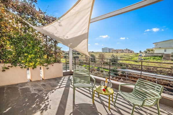 A balcony with a table and chairs, a bottle of champagne, and a view of a green landscape and buildings.