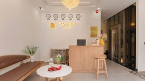 Et hotellets receptionområde med en receptionsdisk, en bænk, et rundt bord med en vase blomster og et ur, der viser tiden i flere byer.