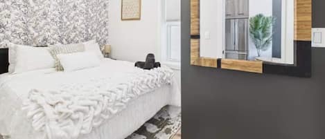A bedroom with a bed, a patterned wall, a framed picture, and a window with a view of a refrigerator and a plant.