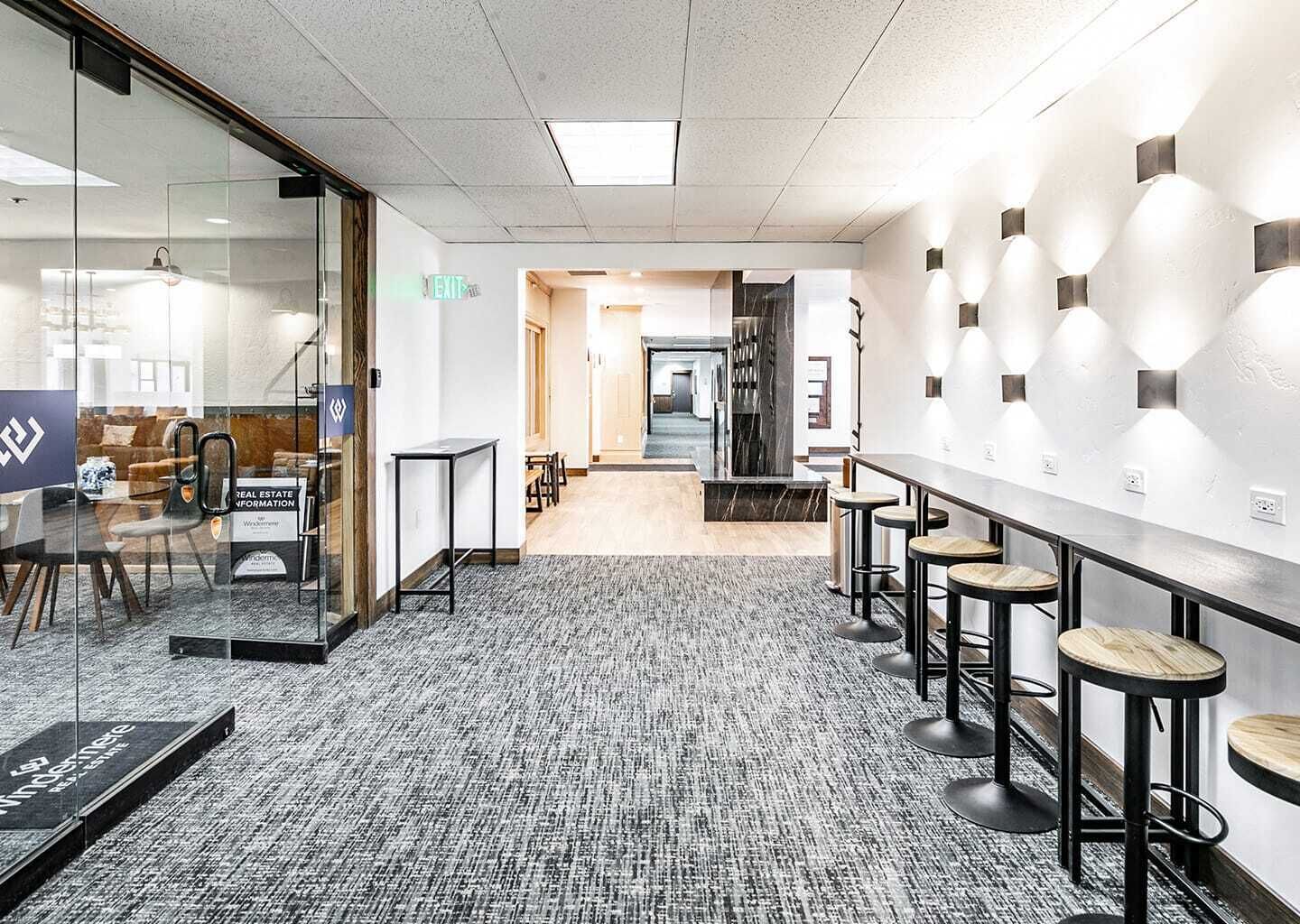 A modern office space with glass walls, a long counter with stools, and a carpeted floor.