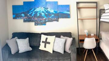 A room with a grey sofa, a small wooden desk, a white chair, and a bunk bed.