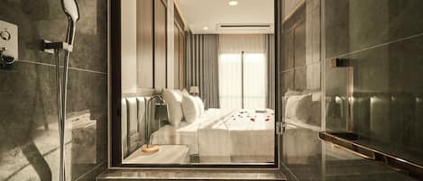 A modern bathroom with a glass shower enclosure, a bed with white linens, and a view of a bedroom with a large window.