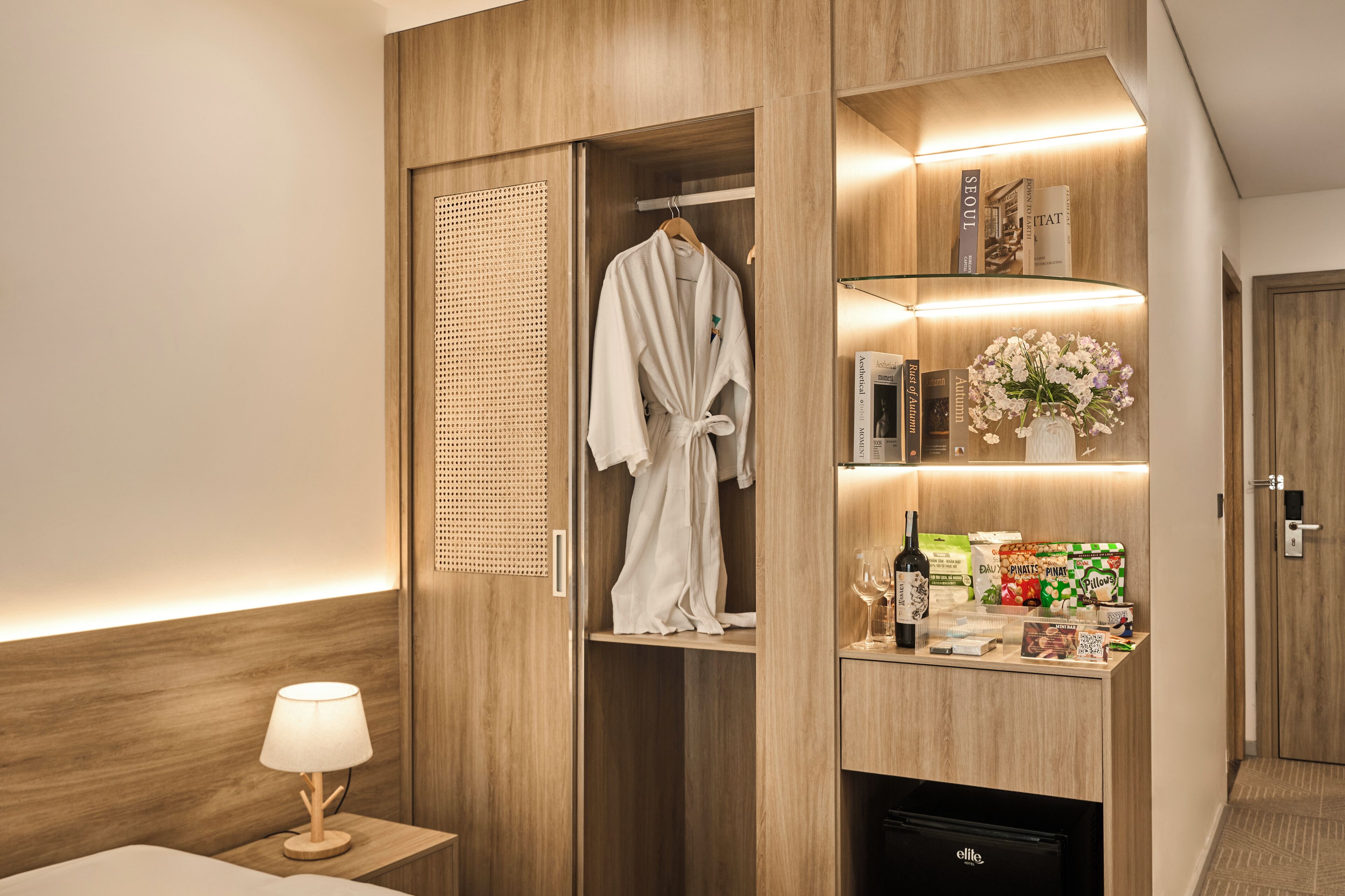 A hotel room with a wooden wardrobe, a robe hanging inside, a bedside table with a lamp, and a minibar with various items.