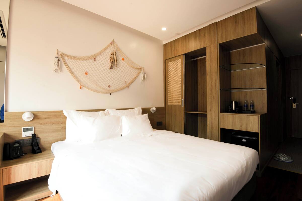 Habitación de hotel con cama, mesita de noche con teléfono y lámpara, armario de madera y estante con artículos de aseo.