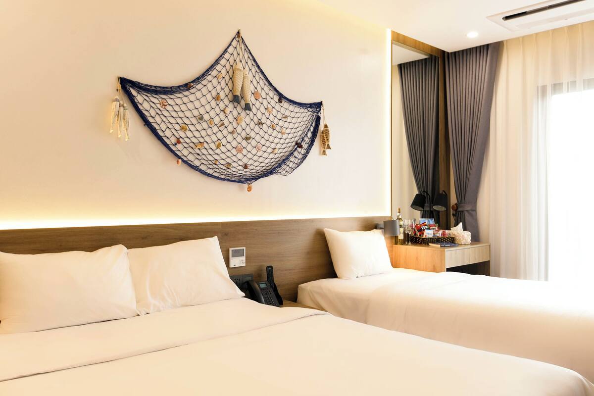 Habitación de hotel con dos camas, teléfono en la pared, mesita de noche con lámpara y una ventana grande con cortinas.