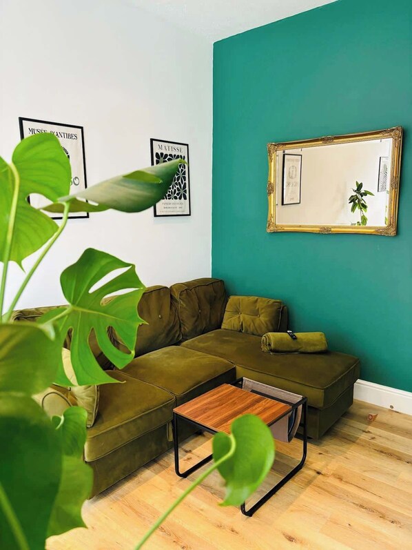 Una sala de estar moderna con un sofá seccional verde, una mesa de centro de madera y un espejo grande en una pared azul verdoso.