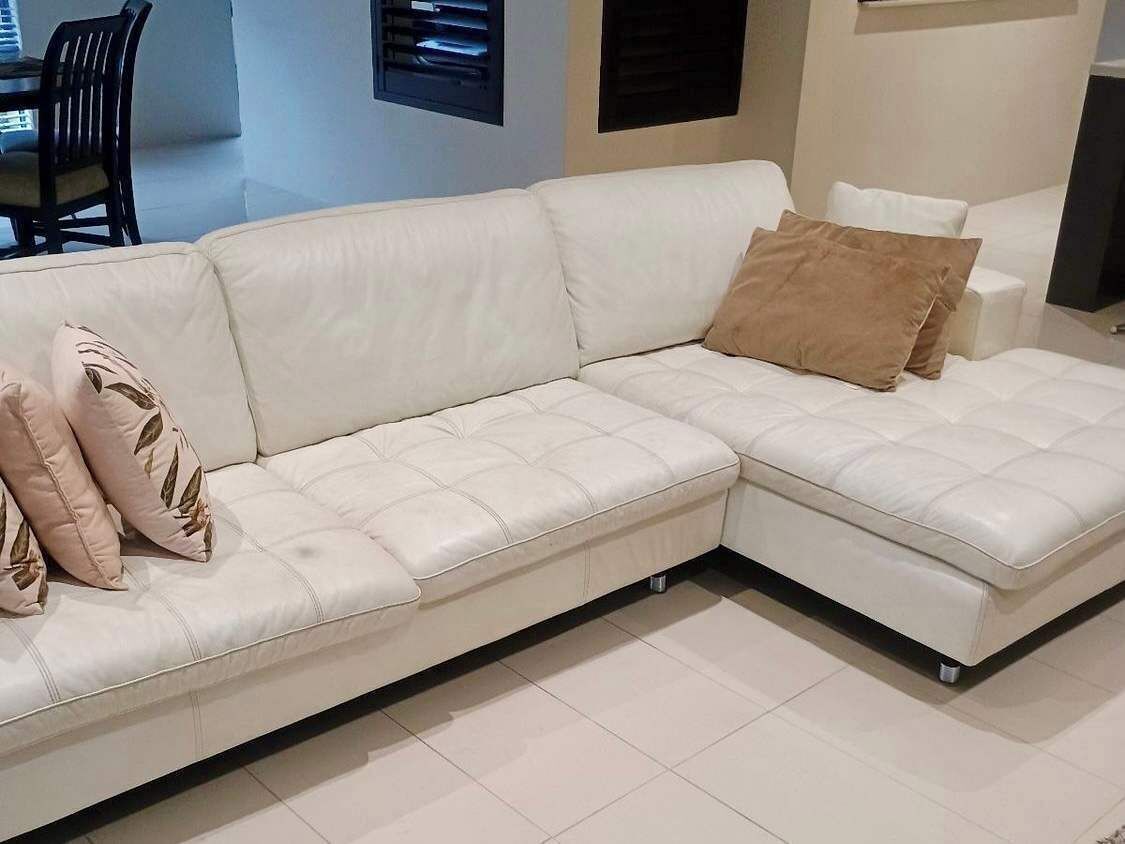 Un sofá seccional de cuero blanco con un cojín marrón, una mesa de comedor con sillas y un aire acondicionado instalado en la pared.