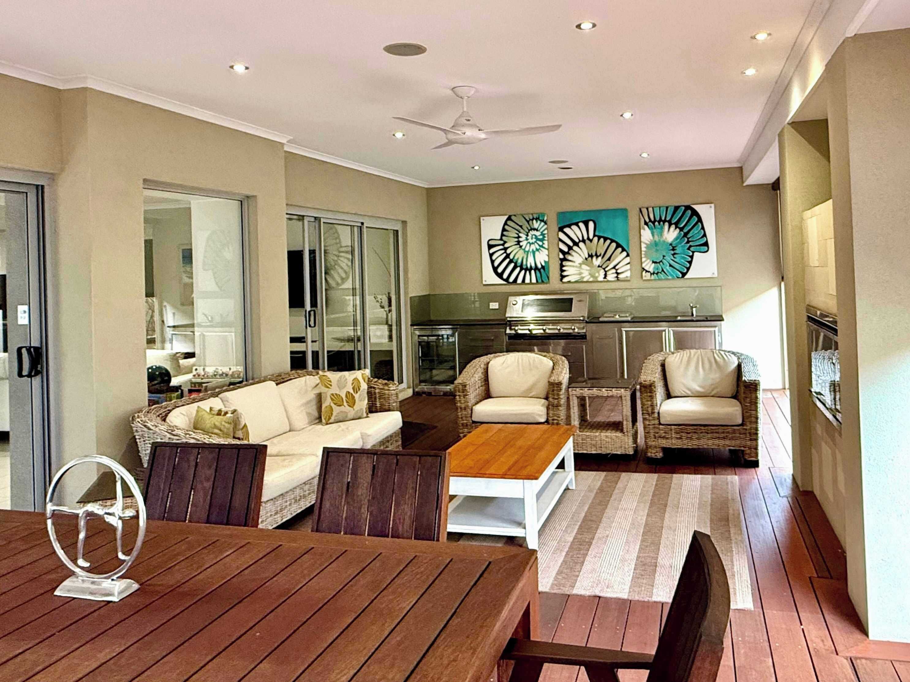 Relax in this covered outside patio area equipped with a lounge couch and dining set. Features stylish wall art, recessed lighting, and sliding doors connecting to the indoor living space.