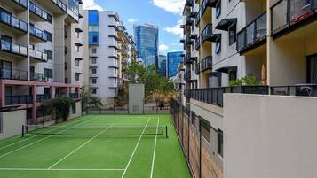 A tennis court surrounded by apartment buildings with balconies, a clear sky, and a modern building in the background.