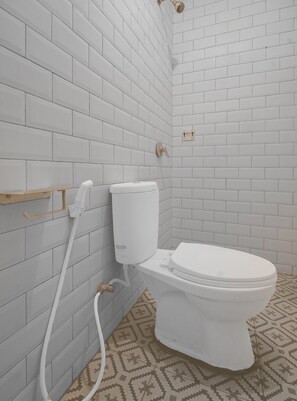 Un baño con paredes de azulejos blancos, un inodoro, un cabezal de ducha y un piso con diseño.