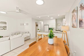 Ein modernes Wohnzimmer mit einem Holzesstisch, einem weißen Sofa und einer Küche im Hintergrund.