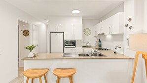Una cocina moderna con armarios blancos, microondas integrado, refrigerador y pileta. Hay dos taburetes de madera en la barra central.
