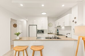 Eine moderne Küche mit weißen Schränken, einer eingebauten Mikrowelle, einem Kühlschrank und einem Spülbecken. An der Kücheninsel stehen zwei Holzschelle.